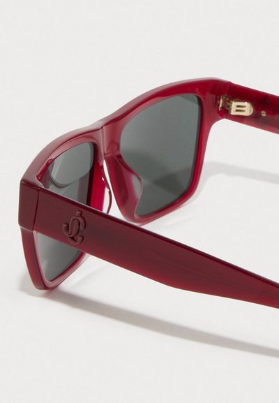 Κόκκινες oversized ηλιόφρες με γυαλιστερό πλαστικό σκελετό, σκούρους φακούς και χρυσή λεπτομέρεια στη μεντεσέ. Διακριτό λογότυπο στο βραχίονες.