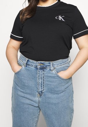 Kvinde iført sort Calvin Klein t-shirt med hvid kant og lyseblå jeans med høj talje, hænderne i lommerne, foran en ensfarvet hvid baggrund.