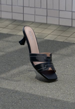 Mule à talon noir à bout ouvert avec double brides et bout carré sur un pavé texturé devant un mur carrelé.