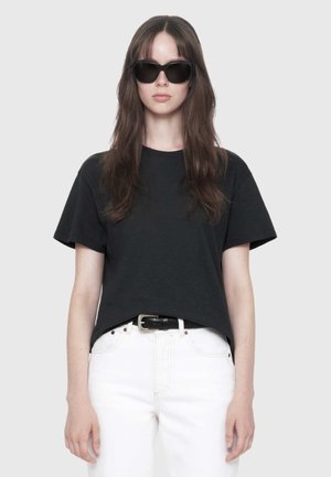 Young woman with long brown hair wearing black sunglasses, a black t-shirt, white pants, and a black belt standing against a plain background.
