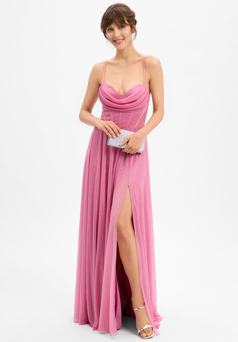 Rosa Abendkleid mit einem Wasserfallausschnitt, schlanken Trägern und einem seitlichen Schlitz. Hergestellt aus schimmerndem Stoff, kombiniert mit einer silbernen Clutch und Sandalen.