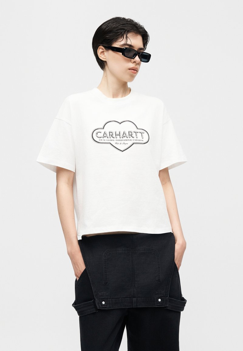 Person trägt weißes Carhartt-T-Shirt mit Logo in Herzform, schwarze Hose, schwarzen Jeansrock um die Taille gebunden und schwarze Sonnenbrille.