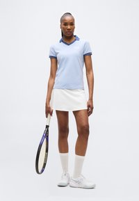 Γυναίκα με ανοιχτό γαλάζιο πουλόβερ πόλο και λευκή φούστα κρατώντας ρακέτα τένις, στέκεται μπροστά σε λευκό φόντο.