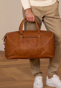 Bruin lederen reistas met dubbele handgrepen, een ritsvak aan de voorkant en contrasterende stiksels. De afmetingen zijn compact en geschikt voor reizen.