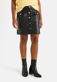 Minifalda de cuero negra con cintura alta, botones a presión en el frente y dos bolsillos frontales. Combinada con botines negros.