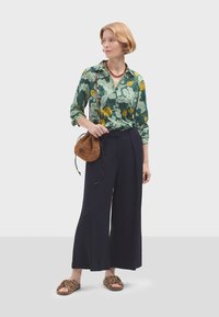 Camicia floreale verde, pantaloni larghi navy, borsa a tracolla intrecciata marrone e sandali a fantasia. Presenta bottoni e un tessuto testurizzato.