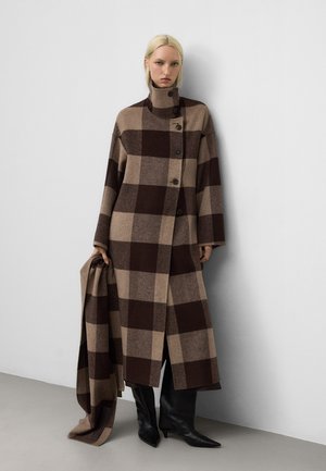 Cappotto lungo in lana con motivo a scacchi marrone e beige, dotato di un colletto alto, chiusura a bottoni e maniche larghe, abbinato a stivali neri a punta.