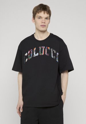 Junger Mann mit kurzen braunen Haaren, trägt ein lockeres schwarzes T-Shirt mit buntem "COLUCCI"-Schriftzug auf der Brust, steht vor einem schlichten Hintergrund.