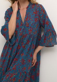 Robe sarcelle avec un col en V et un col volanté, arborant un motif floral en rouge et vert, manches longues évasées et jupe à volants.