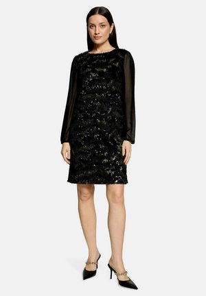 Femme portant une robe noire longueur genou avec des sequins et des manches longues transparentes, associée à des mules noires à talons hauts avec un détail chaîne dorée.