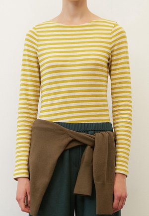 Person trägt ein gelb-weiß gestreiftes Langarmshirt, dunkle Hosen und einen braunen Pullover, der um die Taille gebunden ist.