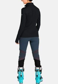Zwarte longsleeve top met hoge kraag, gecombineerd met grijs en zwarte broek met bordeauxrode accenten en turkoois skiboots.