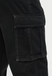 Τσέπη μαύρου παντελονιού cargo με καπάκι και εμφανή λευκή ραφή σε βαριά ύφανση, εμφανιζόμενη στην περιοχή του άνω μηρού.