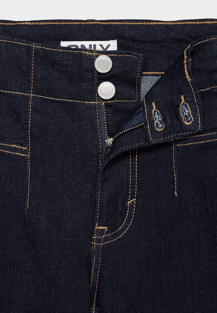 Jean en denim bleu foncé avec une taille haute, deux boutons en argent et des coutures dorées contrastantes. La fermeture éclair est partiellement visible.