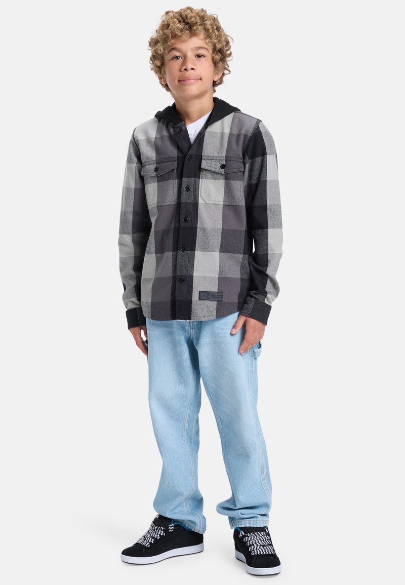 Černá a šedá kostkovaná flanelová košile s černou kapucí, dvěma kapsami na hrudi, ve spojení se světle modrými džíny a černými teniskami s bílými akcenty.