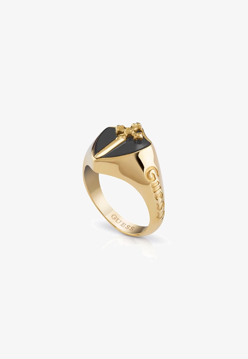 Gold ring with a black shield-shaped stone featuring a decorative cross design. "Guess" logo engraved on the band. Shiny, smooth texture.