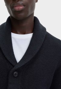 Donkerblauwe cardigan met een gestructureerd breipatroon, voorzien van een kraag en een enkele zwarte knoop. Draagt over een wit T-shirt.