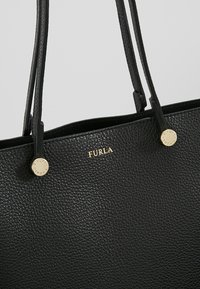 Sac à main Furla en cuir noir texturé avec deux poignées fines doubles et détails logo et boutons dorés sur un fond gris clair.