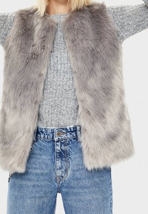 Person iført en grå faux pelsvest over en grå strikket sweater med højtaljede blå jeans, armene let løftet uden for billedets ramme.