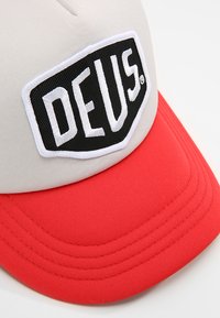 Gorra de béisbol con una corona beige, visera roja y un parche rectangular negro con el texto blanco "DEUS". Textura suave, forma estructurada.
