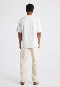 Hombre con cabello rizado corto de espaldas, vistiendo una camisa blanca de manga corta, pantalones color crema y zapatillas blancas con suelas marrones.