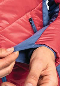 Rote gesteppte Jacke mit blauen Innenbündchen, ausgestattet mit einer Reißverschlusstasche an der Seite und weichem Futter, das aus dem geöffneten Bündchenbereich sichtbar ist.