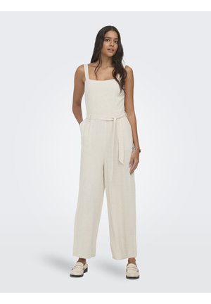 Femme debout, les mains dans les poches, portant une combinaison sans manches couleur crème avec une ceinture et des mocassins assortis sur un fond uni.