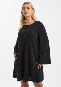 Musta mekko pitkillä puffimaisilla hihoilla, jossa on teksturoitu kangas, jossa on pieniä valkoisia pisteitä. Flare-mallinen siluetti, pyöreä pääntie.