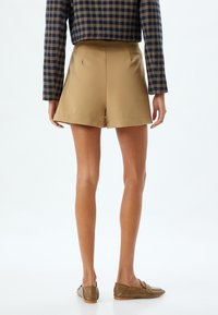Pantaloni beige su misura con una texture liscia e aperture delle gambe ampie, abbinati a mocassini marroni, visti da dietro con una maglietta a scacchi.
