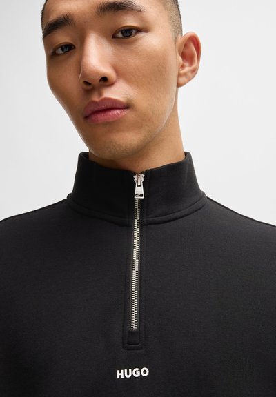 Sweatshirt noir à fermeture éclair avec col haut, tissu texturé, fermeture éclair argentée et logo "HUGO" blanc sur la poitrine. Coupe structurée.