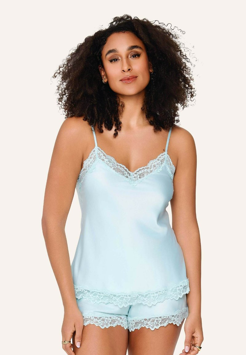 Femme aux cheveux bouclés portant un caraco bleu clair orné de dentelle et un short, debout devant un fond beige uni.