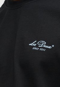 Μαύρο φούτερ με στρογγυλή λαιμόκοψη. Διαθέτει λευκό κεντητό κείμενο που γράφει "Les Deux since 2011." Ομαλή υφή και απλός σχεδιασμός.