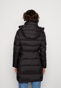 Manteau matelassé noir avec capuche, présentant un design matelassé, des manches longues et une coupe jusqu'aux genoux. Tissu lisse et brillant.