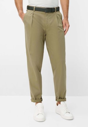 Mand iført kaki bukser med op-rullede manchetter, hvide sneakers, sort bælte og hvid skjorte, står med hænderne i lommerne.