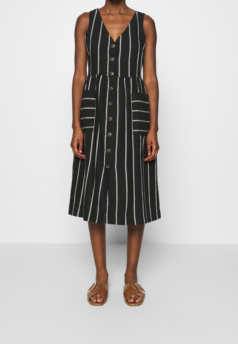 Robe sans manches à rayures noir et blanc avec un col en V, devant à boutons, deux poches avant et une longueur de genoux. Portée avec des sandales marron.