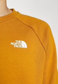 Sudadera de manga larga en color mostaza, hecha de tela texturizada. Presenta un logo blanco en el pecho y un cuello y puños acanalados.