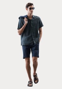 Kurzärmliges, dunkelgrünes Leinenhemd, marineblaue Shorts, braune Sandalen mit Riemen und schwarze Sonnenbrille. Das Modell trägt einen marineblauen Pullover über der Schulter.