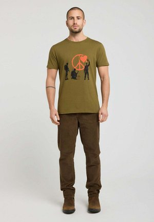 Un homme se tient face à nous portant un t-shirt vert olive avec un symbole de paix rouge et un dessin de cœur, ainsi qu’un pantalon marron et des chaussures marron.