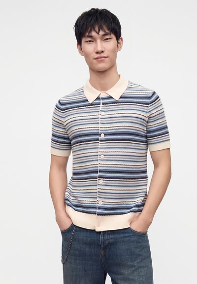 Jeune homme portant une chemise rayée à manches courtes boutonnée avec un col et des poignets beiges, debout les mains dans les poches d'un jean bleu.