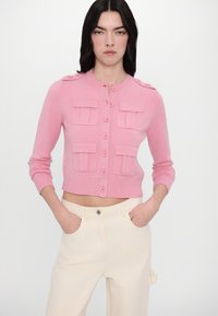 Light pink cropped cardigan with four front pockets, round neckline, and button closure. Knit texture with ribbed accents at cuffs and hem.