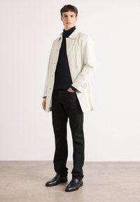 Abrigo color crema con diseño de cuello, suéter negro de cuello alto, pantalones negros ajustados y zapatos negros. Textura suave, estilo minimalista.