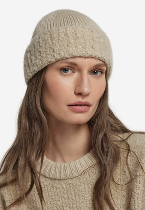Beige, strukturierte Strickmütze mit geripptem oberem Abschnitt; weiche Wolle und runde Form, getragen von einem Modell mit langen Haaren und neutralfarbener Kleidung.