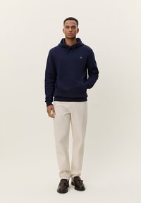 Tmavomodrá mikina s kapucňou z mäkkého materiálu s klokaním vreckom a šnúrkou na kapucni, skombinovaná so svetlobéžovými nohavicami a tmavohnedou obuvou.