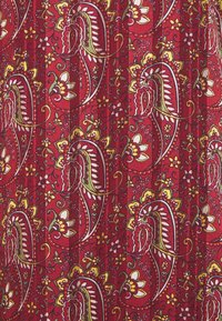 Punainen kangas, jossa on monimutkainen paisley-kuvio keltaisena, valkoisena ja vihreänä. Sisältää kukkaaksentteja ja tekstuurin näköisen pinnan.