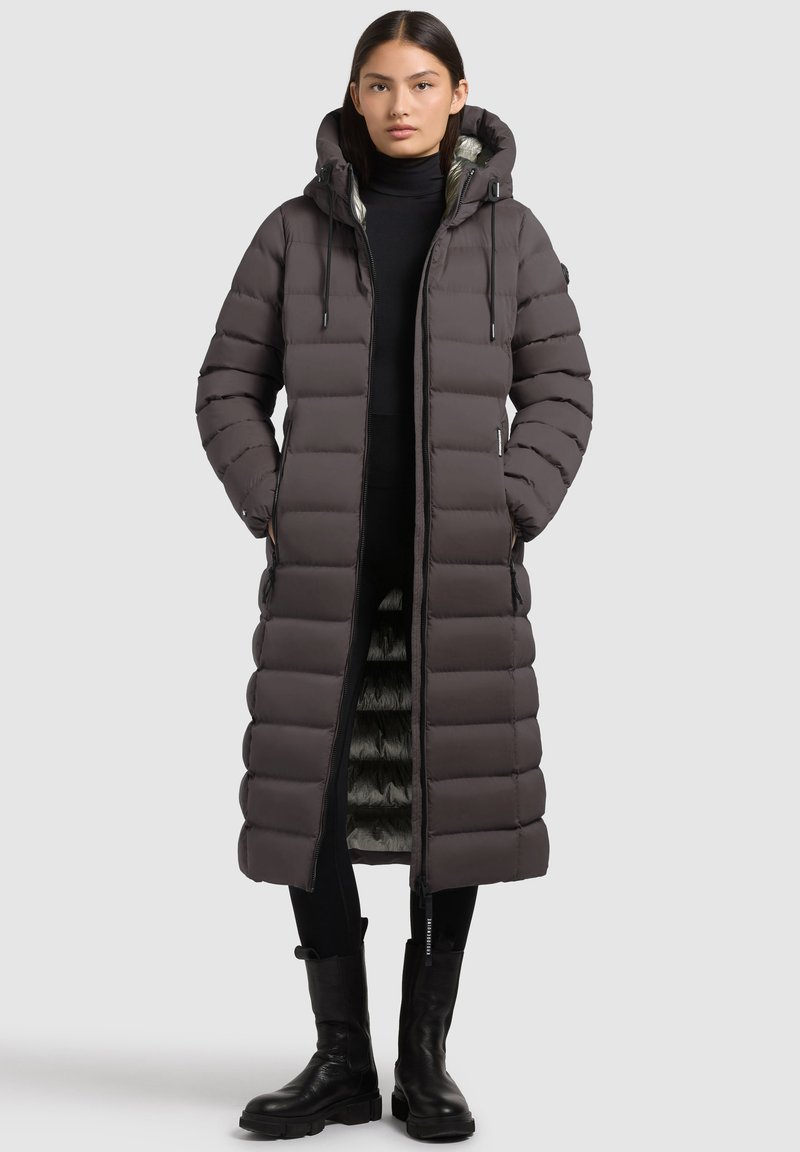 Cappotto lungo, con cappuccio, imbottito di un grigio scuro con interni lucidi. Presenta una cerniera frontale, tasche laterali e polsini a coste. Indossato sopra un abbigliamento nero.