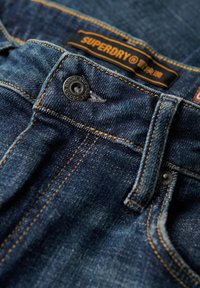 Ciemnoniebieskie dżinsy z denimu z pomarańczowym przeszyciem, metalowym guzikiem z wygrawerowanym logo oraz czarną metką z napisem "SUPERDRY" na pasku talii.