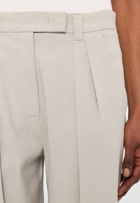 Lichtgrijze broek met scherp geplooide details, voorzien van een gladde textuur en een aansluitende tailleband. Minimalistisch ontwerp zonder zichtbare hardware.