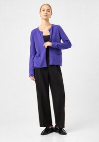 Kvinde iført en lilla knap-op cardigan over en sort top, sorte vidde bukser og sorte loafers, stående foran en ensfarvet baggrund.