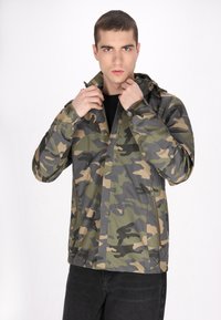 Vihreä-beige sävyinen maastopuku, valmistettu kevyestä materiaalista, jossa on huppu, vetoketjulliset taskut ja säädettävät hihansuut.