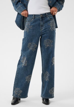 Personne portant un jean bleu à jambes larges avec des motifs de roses argentées, associé à des bottes noires et un haut blanc, mains ajustant les côtés de la veste.